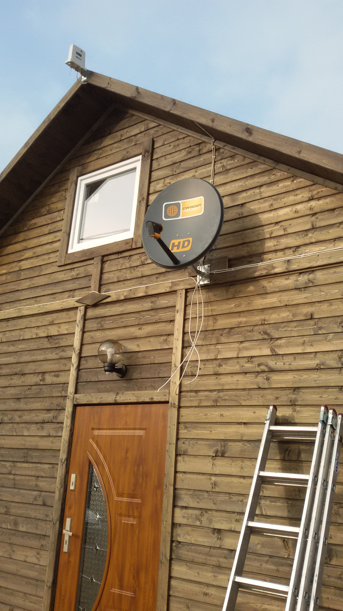 Antena satelitarna 'CYFROWY' HD zamontowana na drewnianej elewacji domu, widoczne wystające kable, lampa zewnętrzna i drabina oparta o ścianę.