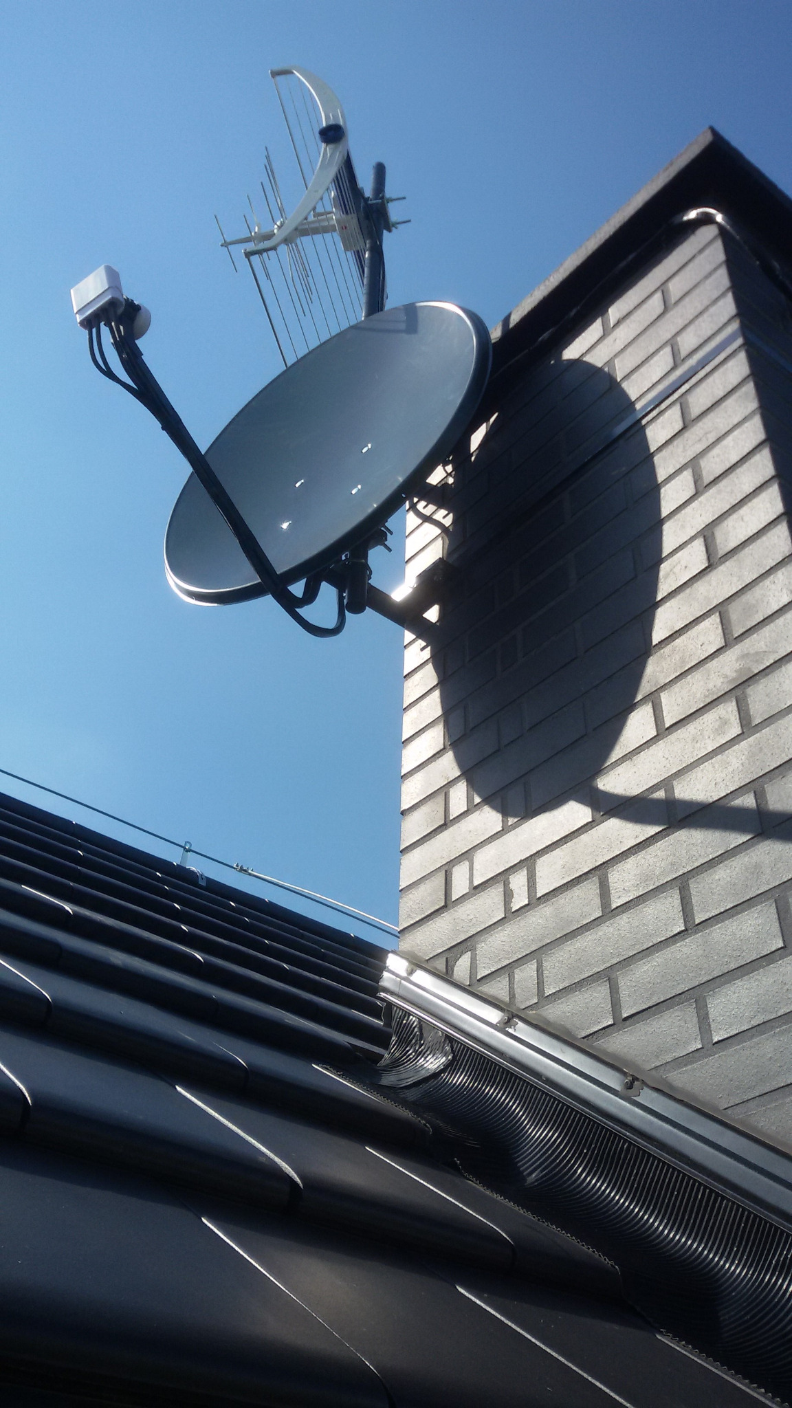 Antena satelitarna i antena naziemna zamontowane na kominie z cegły, widok z dołu na tle błękitnego nieba.