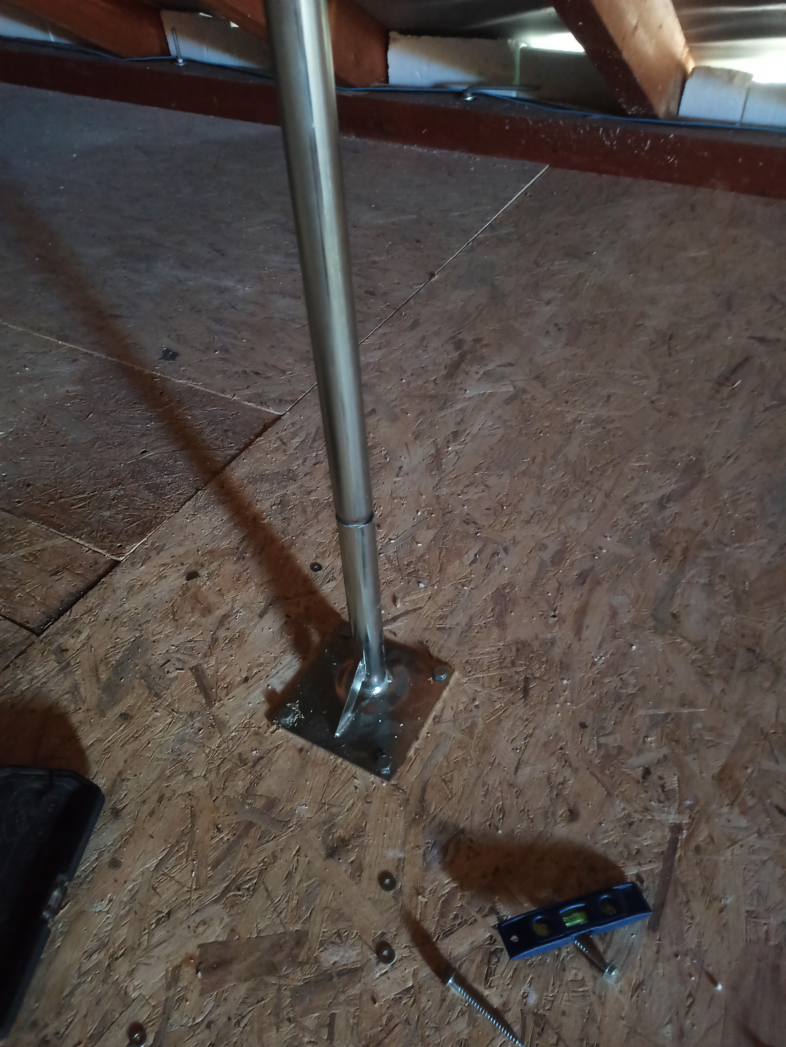 Metalowy maszt antenowy zamontowany na drewnianej podłodze strychu, poziomica leży obok.