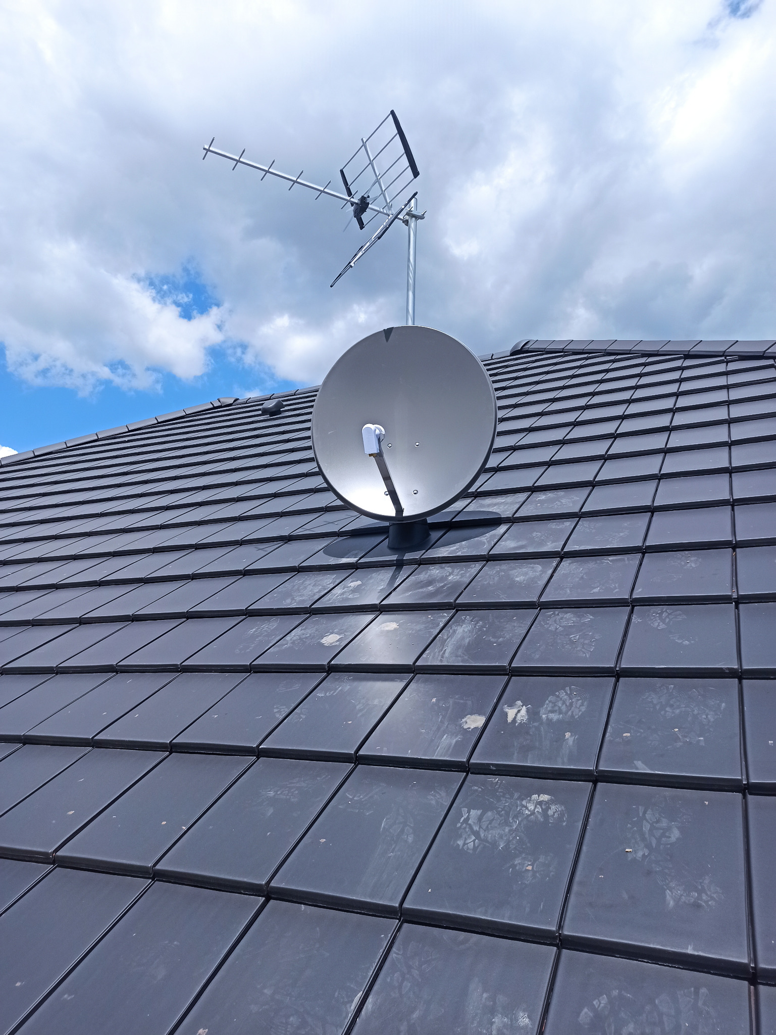 Antena satelitarna i antena naziemna zamontowane na ciemnoszarym dachu z dachówki ceramicznej, widok z dołu na tle błękitnego nieba z białymi chmurami.