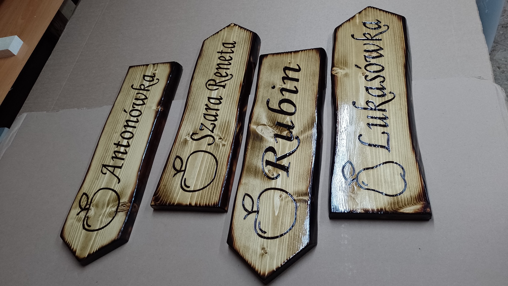 Ręcznie robione, wypalane deski z nazwami odmian jabłek (Antonówka, Szara Reneta) i gruszek (Rubin, Łukasówka), drewno opalane, lakierowane, widoczne słoje.