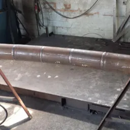 Długa, metalowa rura z widocznymi spawami, umieszczona na metalowych stojakach w warsztacie; na rurze widoczne białe oznaczenia.