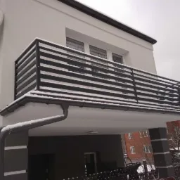 Nowoczesna, pozioma balustrada balkonowa z czarnych profili, pokryta śniegiem, na tle białej elewacji budynku z anteną satelitarną.
