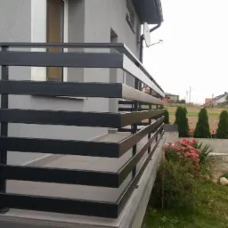 Nowoczesna balustrada balkonowa z poziomych, czarnych profili, zamontowana na jasnoszarym budynku z widoczną anteną satelitarną i zielonym ogrodem z tujami w tle.