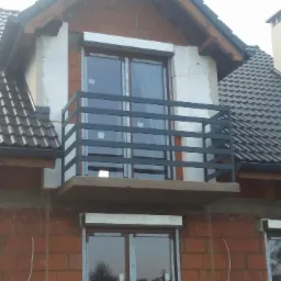 Szara, metalowa balustrada balkonowa z poziomymi szczeblami, zamontowana na budynku w trakcie budowy, z widocznymi elementami konstrukcyjnymi i oknami z roletami.