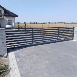 Nowoczesna brama wjazdowa przesuwna z poziomych profili, szara kostka brukowa, słupki z szarego betonu architektonicznego, dom jednorodzinny w tle, widok z zewnątrz posesji.