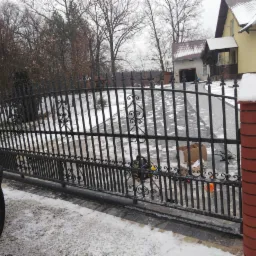 Czarna brama wjazdowa z kutymi zdobieniami, częściowo zamontowana, widoczne narzędzia i kartonowe pudło, na tle domu z kostki brukowej i zimowego krajobrazu.