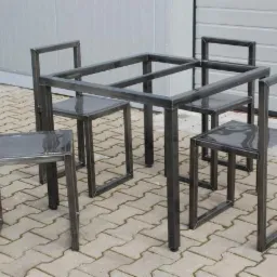 Metalowy stół z czterema krzesłami o minimalistycznej, surowej konstrukcji z profili stalowych, ustawiony na kostce brukowej przed szarą, metalową ścianą.