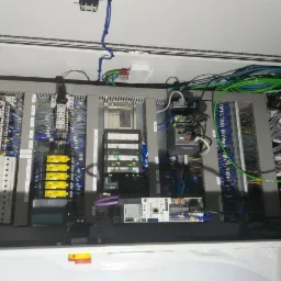 Otwarta szafa sterownicza z widocznymi modułami, przewodami i złączami, uporządkowanymi wewnątrz obudowy, z elementami oznaczonymi kolorami.