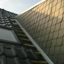 Fragment dachu z czarną dachówką ceramiczną i szarym łupkiem, widoczna metalowa rynna i okno dachowe.