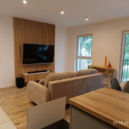 Jasne wnętrze salonu z drewnianą ścianą TV, beżową sofą i podłogą imitującą drewno, widok z perspektywy blatu kuchennego.