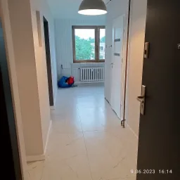 Widok z korytarza na wyremontowane wnętrze mieszkania z białymi marmurowymi płytkami na podłodze, widocznym oknem, grzejnikiem i workiem z narzędziami w tle.