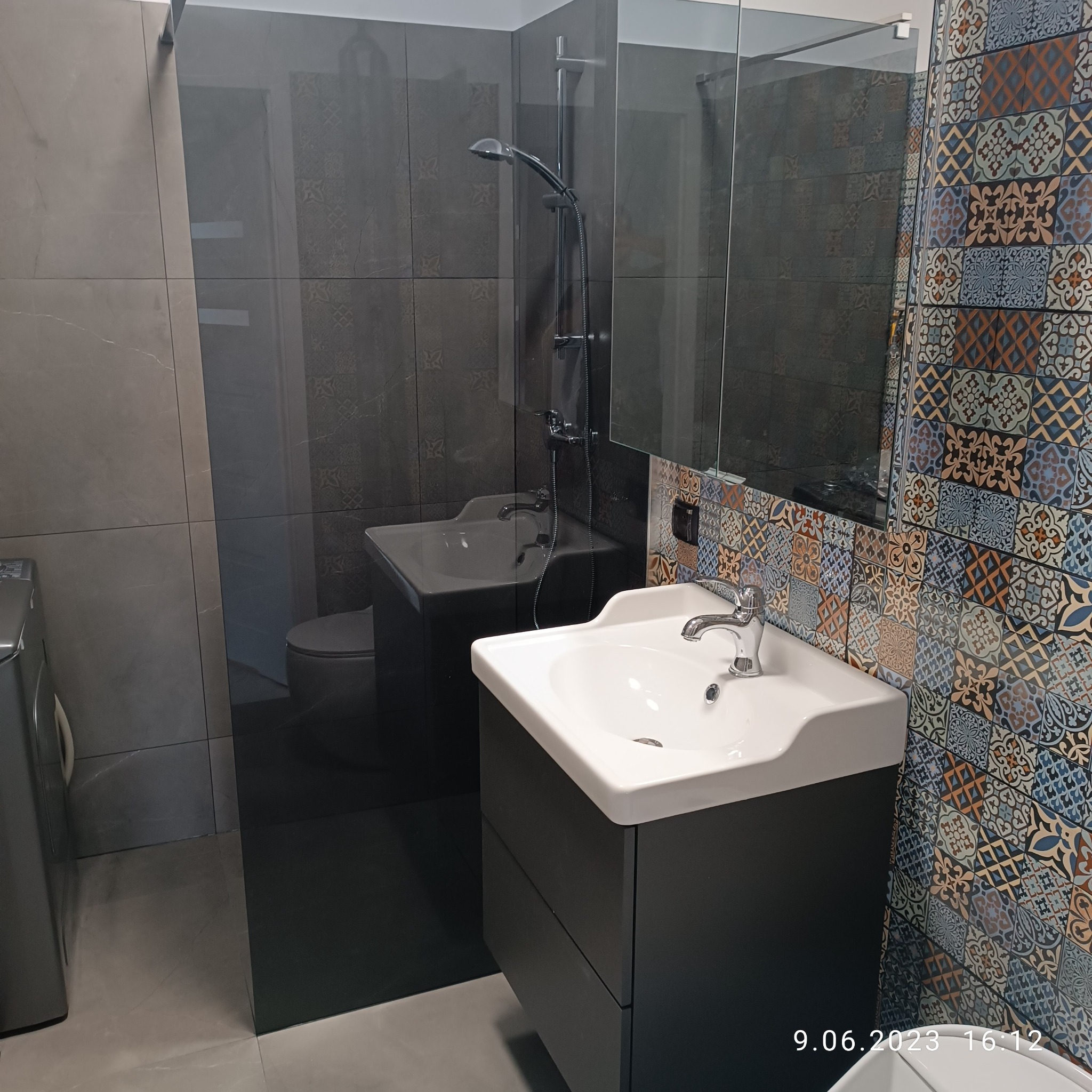 Umywalka z szafką w nowoczesnej łazience z prysznicem typu walk-in i dekoracyjnymi płytkami ściennymi w stylu patchwork, widok na toaletę w lustrze.