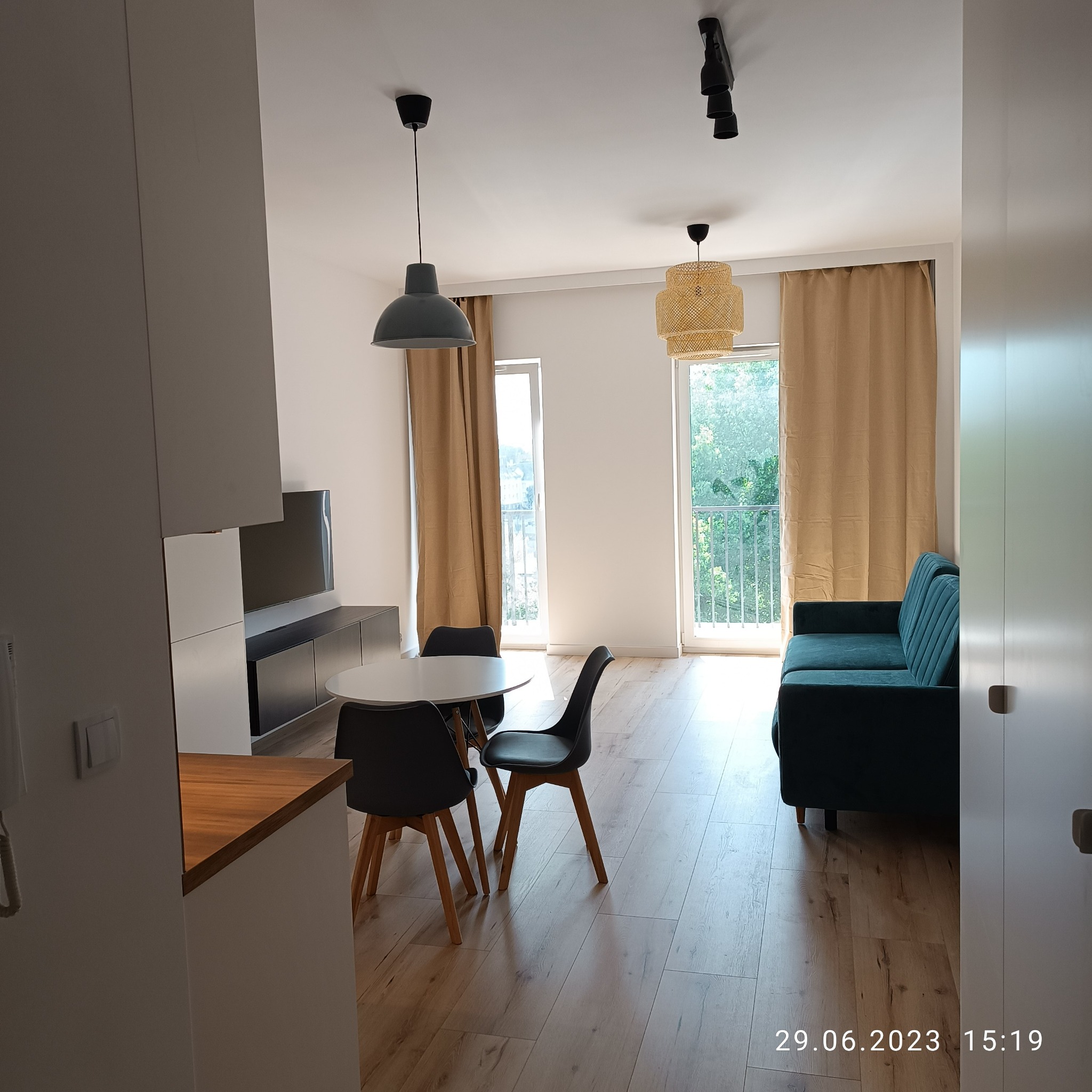 Jasne wnętrze mieszkania po remoncie: okrągły stół z krzesłami, zielona sofa, drewniana podłoga, zasłony w kolorze beżowym i lampy sufitowe.