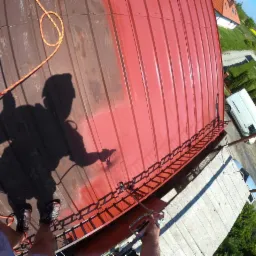 Widok z góry na częściowo pomalowany dach z blachy, cień malarza z pistoletem natryskowym, liny zabezpieczające, w tle zabudowa jednorodzinna i samochody.