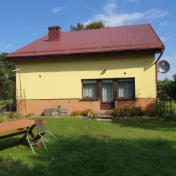 Dom jednorodzinny z nowym, czerwonym dachem z blachy, żółtą elewacją i brązowym cokołem, widoczny z poziomu trawnika.