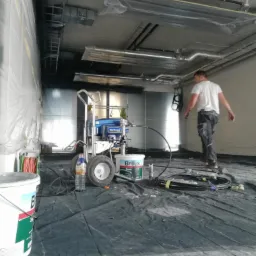 Przygotowane pomieszczenie do malowania natryskowego z maszyną malarską, wiaderkiem farby Brilux i rozłożoną folią ochronną na podłodze, widoczny fragment pomalowanej ściany i pracownik w tle.