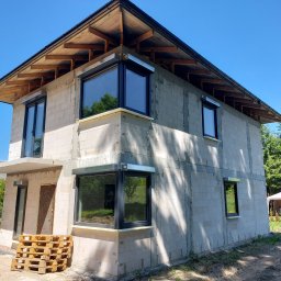 KT - BUD Usługi Budowlane Kamil Turkiewicz - Surowy stan dwukondygnacyjnego domu z betonu komórkowego z zamontowanymi oknami i drewnianą konstrukcją dachu, widoczne palety przed wejściem.