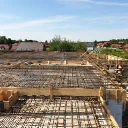 Zbrojenie stropu z widocznymi szalunkami drewnianymi i pionowymi rurami kanalizacyjnymi, w tle zabudowa jednorodzinna i błękitne niebo.