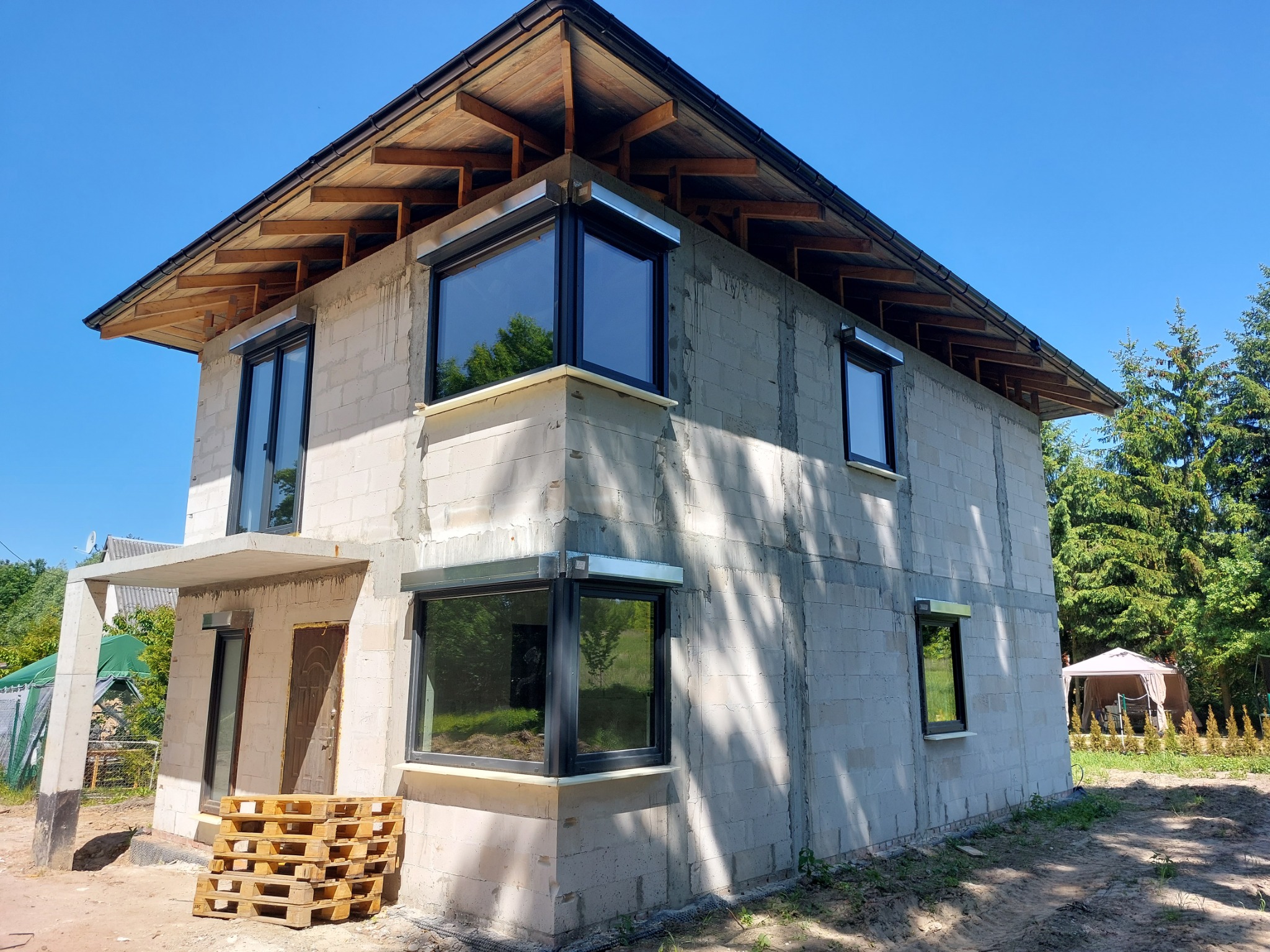 Surowy stan dwukondygnacyjnego domu z betonu komórkowego z zamontowanymi oknami i drewnianą konstrukcją dachu, widoczne palety przed wejściem.