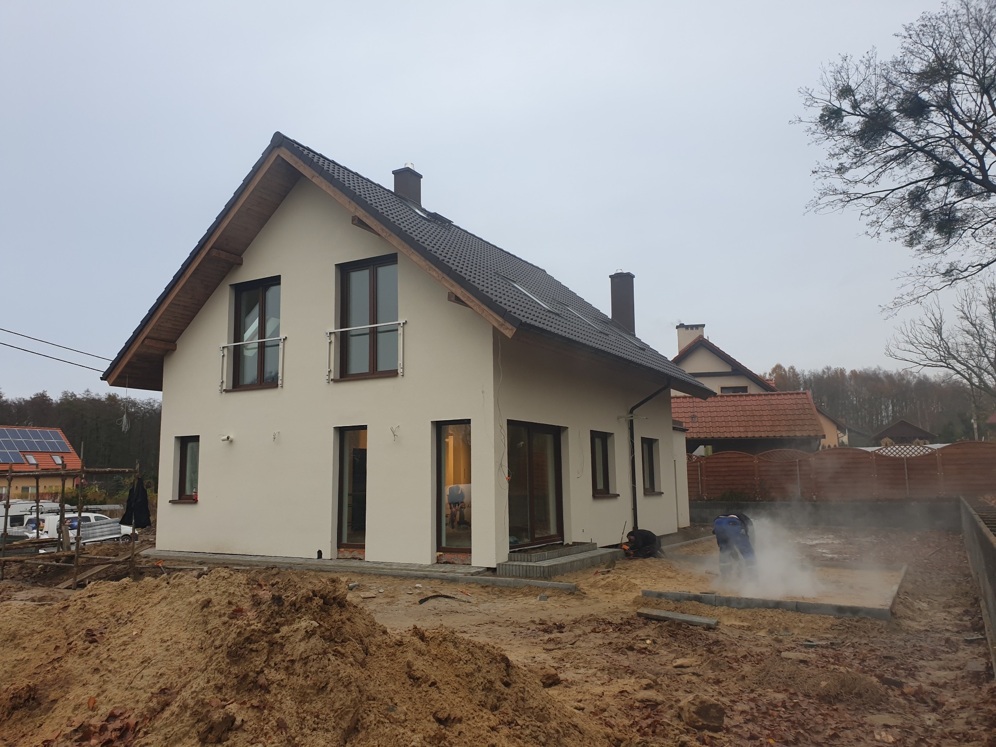 Nowoczesny, dwukondygnacyjny dom z jasną elewacją i ciemną dachówką w trakcie wykańczania; widoczny stos piasku i robotnik układający kostkę brukową przy fundamencie.