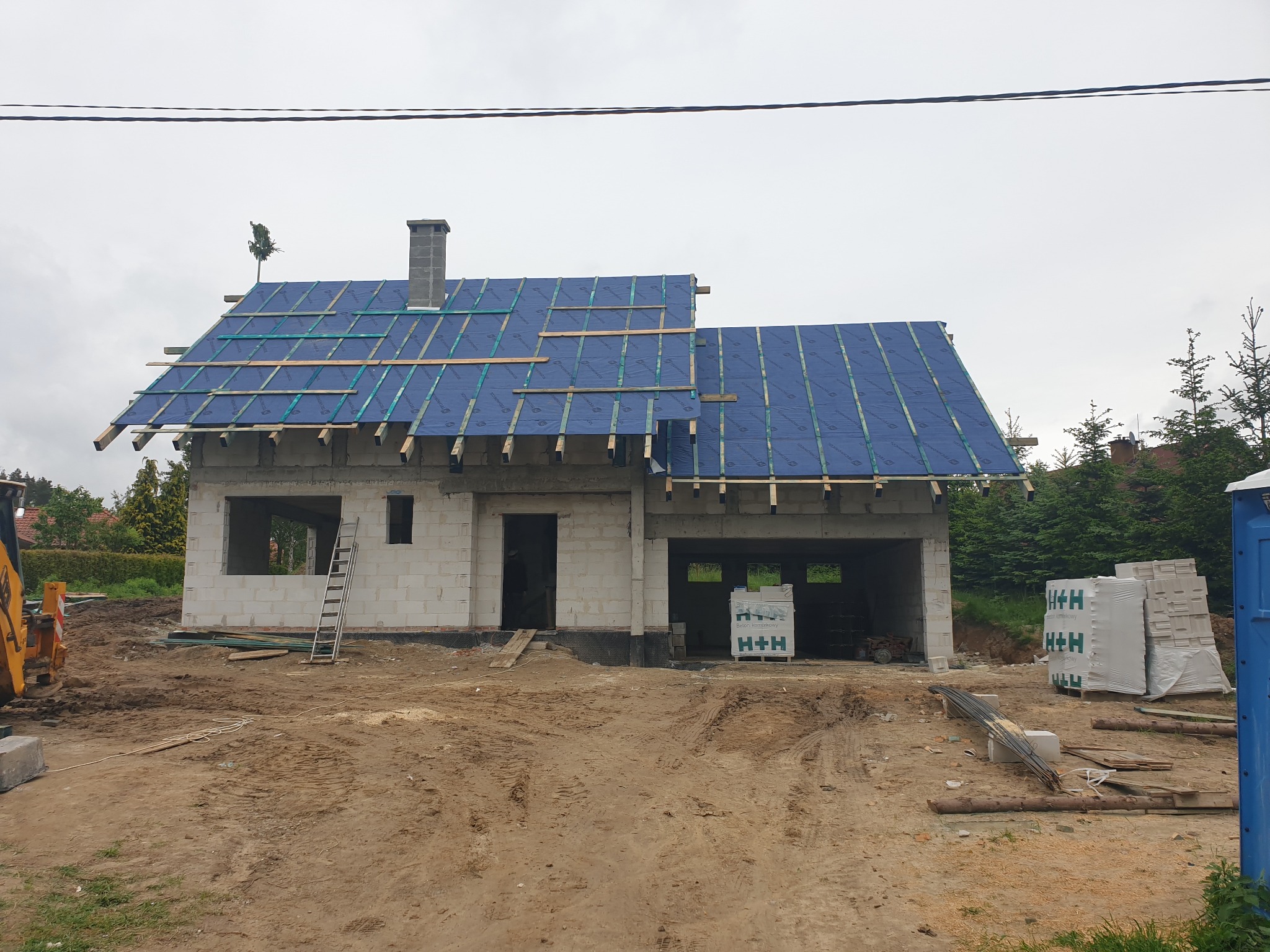 Surowy stan domu jednorodzinnego z betonu komórkowego w trakcie budowy dachu, widoczne krokwie i membrana dachowa, na pierwszym planie teren budowy z ziemią i materiałami budowlanymi.