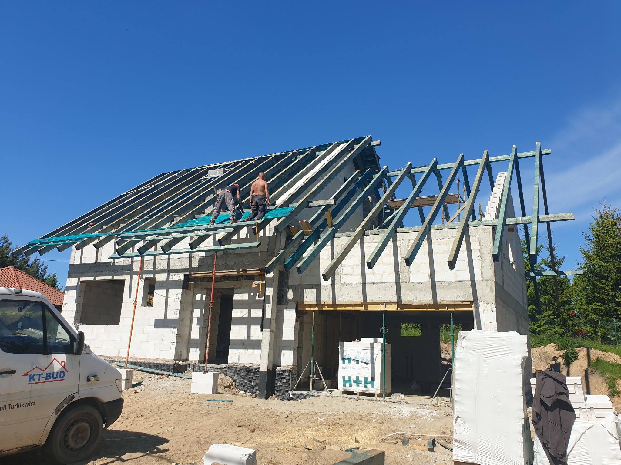 Wznoszenie konstrukcji dachu na surowym budynku z betonu komórkowego, dwóch robotników pracuje na wysokości, widoczny samochód dostawczy z logo firmy budowlanej w tle.