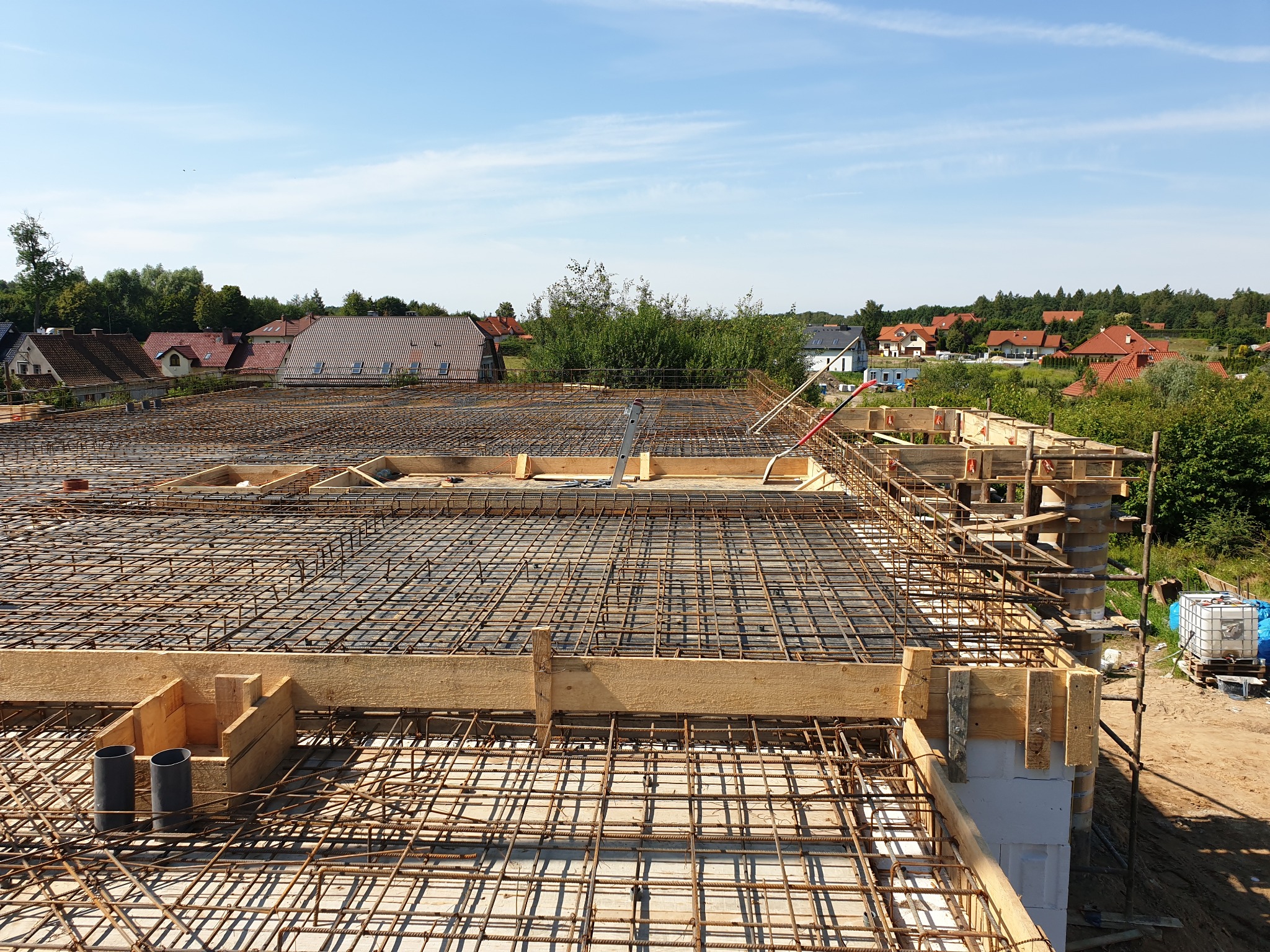 Zbrojenie stropu z widocznymi szalunkami drewnianymi i pionowymi rurami kanalizacyjnymi, w tle zabudowa jednorodzinna i błękitne niebo.