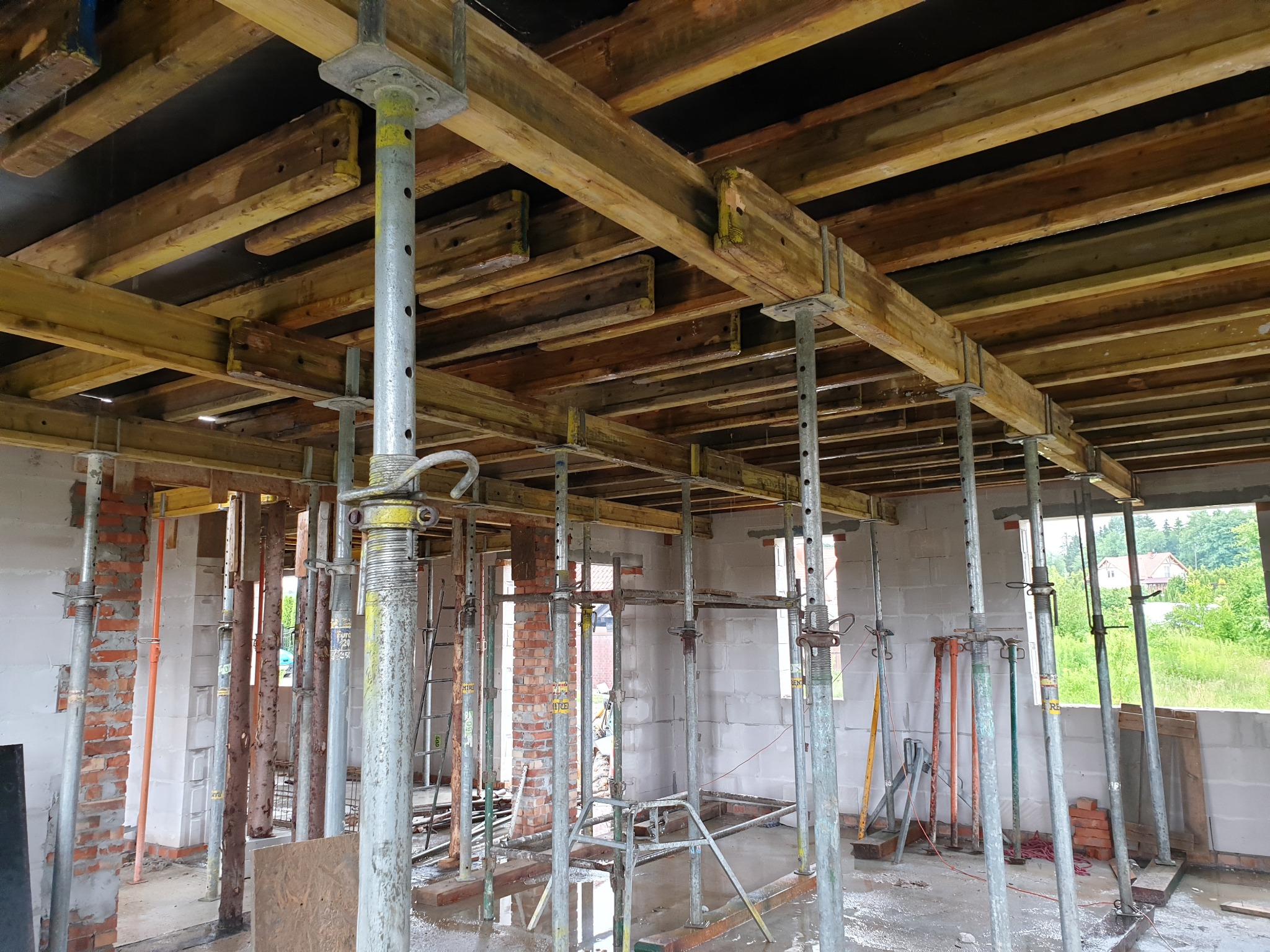 Wnętrze budowanego domu z widocznym szalunkiem stropu podpartym stalowymi stemplami, ściany z bloczków i cegły w surowym stanie, mokra posadzka.