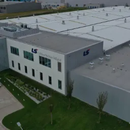 Widok z lotu ptaka na budynek przemysłowy LS Cables System Poland z dachem z płyt warstwowych i elementami wentylacyjnymi, otoczony zielenią i parkingiem.