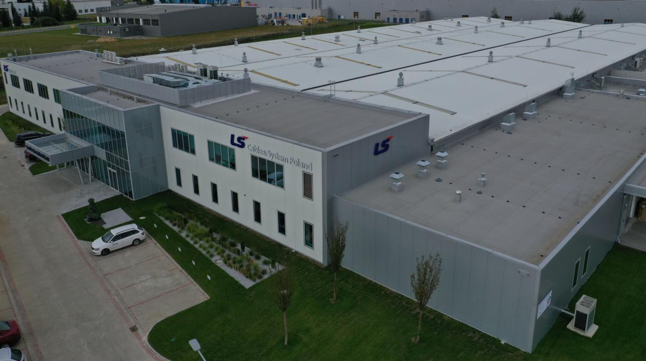 Widok z lotu ptaka na budynek przemysłowy LS Cables System Poland z dachem z płyt warstwowych i elementami wentylacyjnymi, otoczony zielenią i parkingiem.