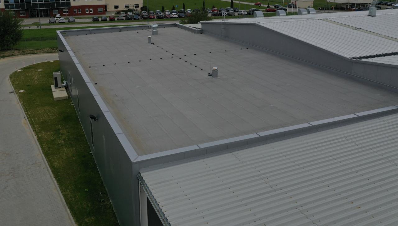 Widok z góry na płaski dach przemysłowy pokryty papą termozgrzewalną z widocznymi kominkami wentylacyjnymi oraz fragment dachu z blachy trapezowej i budynek w tle.