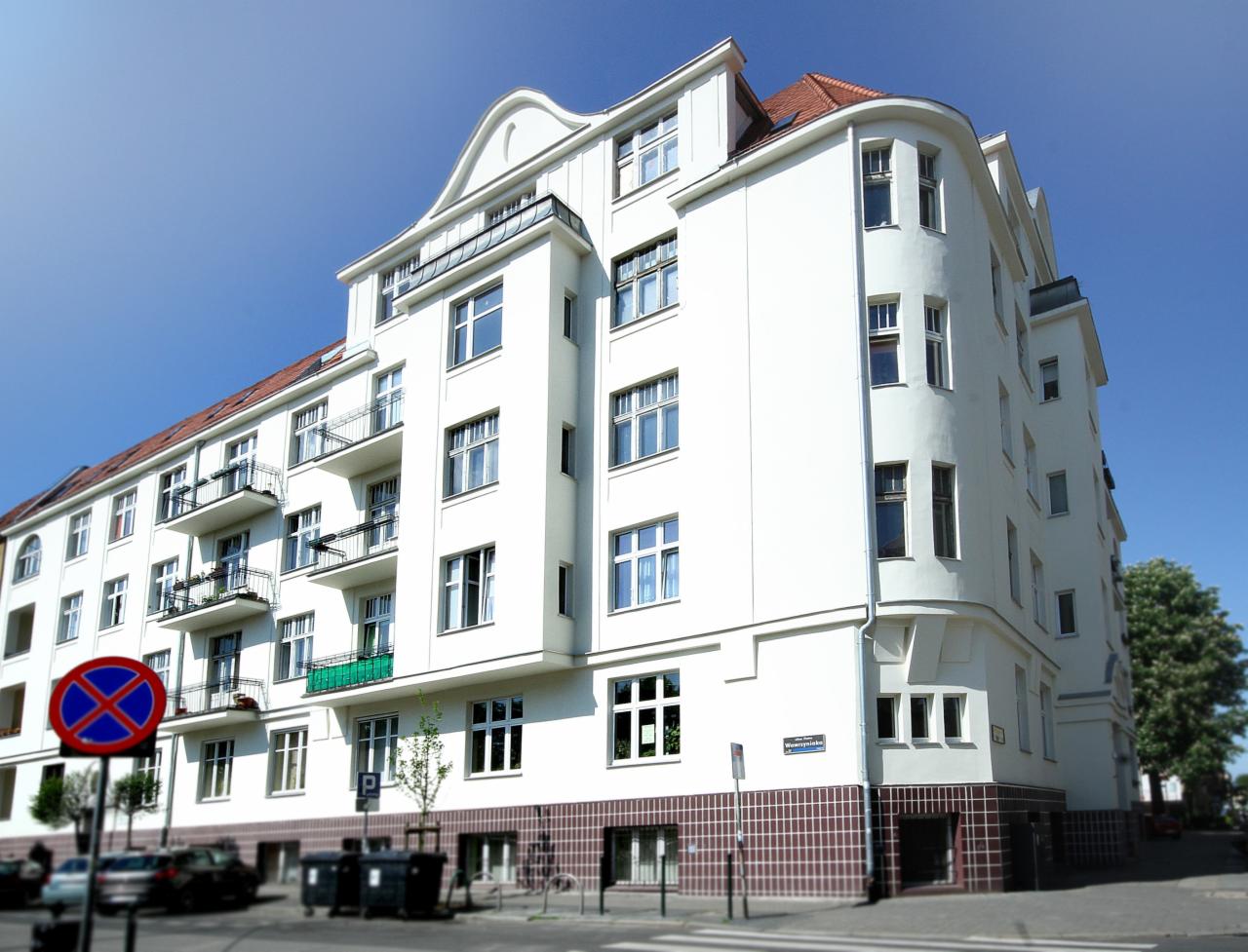Odnowiona kamienica w jasnej kolorystyce z balkonami, widoczna z perspektywy ulicy w słoneczny dzień, z charakterystycznym znakiem drogowym zakazu postoju na pierwszym planie.