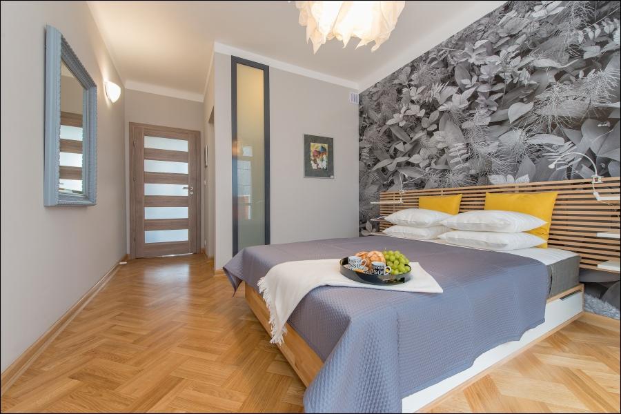 Nowoczesna sypialnia z parkietem w jodełkę, łóżkiem z szarą narzutą, żółtymi poduszkami i dekoracyjną tapetą w motywy roślinne za wezgłowiem, na łóżku taca ze śniadaniem.