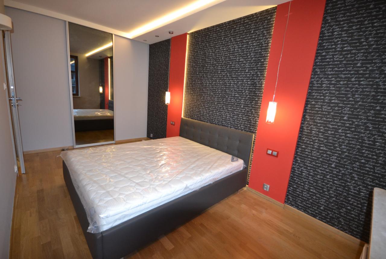 Nowoczesna sypialnia z łóżkiem w szarym obiciu, lustrzaną szafą przesuwną i ścianą w czarno-białe pismo, oświetlona wiszącymi lampami na tle czerwonych pasów.