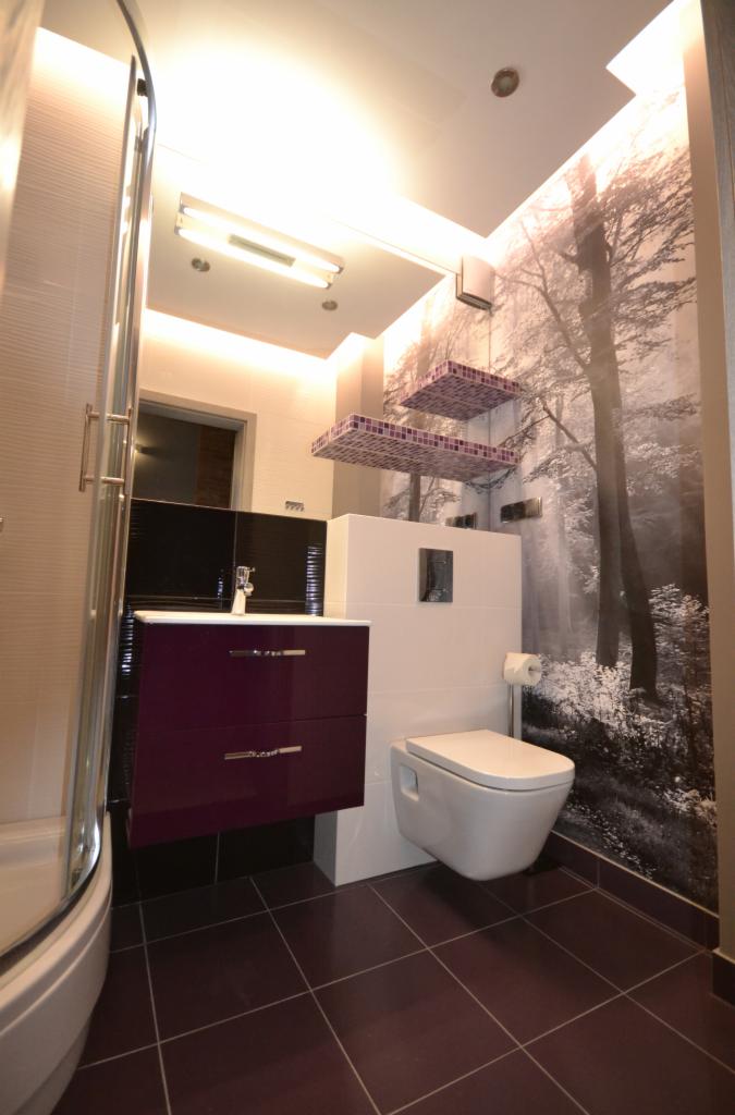 Nowoczesna łazienka z fioletową szafką pod umywalką, wiszącą toaletą i fototapetą z motywem leśnym.