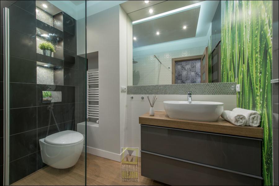 Nowoczesna łazienka z czarnymi płytkami, wnękami z oświetleniem, wiszącą toaletą, drewnianą podłogą i umywalką na drewnianym blacie oraz panelem z motywem trawy na szklanej ściance prysznica.