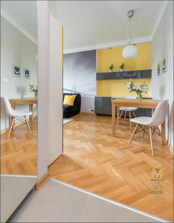 Wnętrze mieszkania z parkietem ułożonym w jodełkę, widok przez otwarte drzwi na salon z żółtą ścianą i fototapetą z mostem, białe krzesła przy drewnianym stole, odbicie w lustrze.