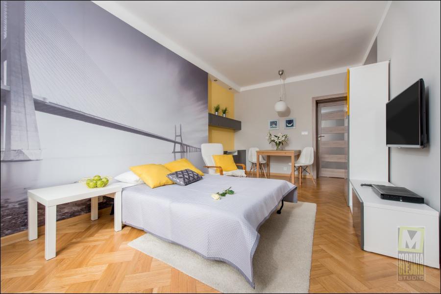 Jasne mieszkanie z sypialnią i aneksem jadalnym, z parkietem w jodełkę, fototapetą z motywem mostu na ścianie, żółtymi akcentami i białymi meblami.