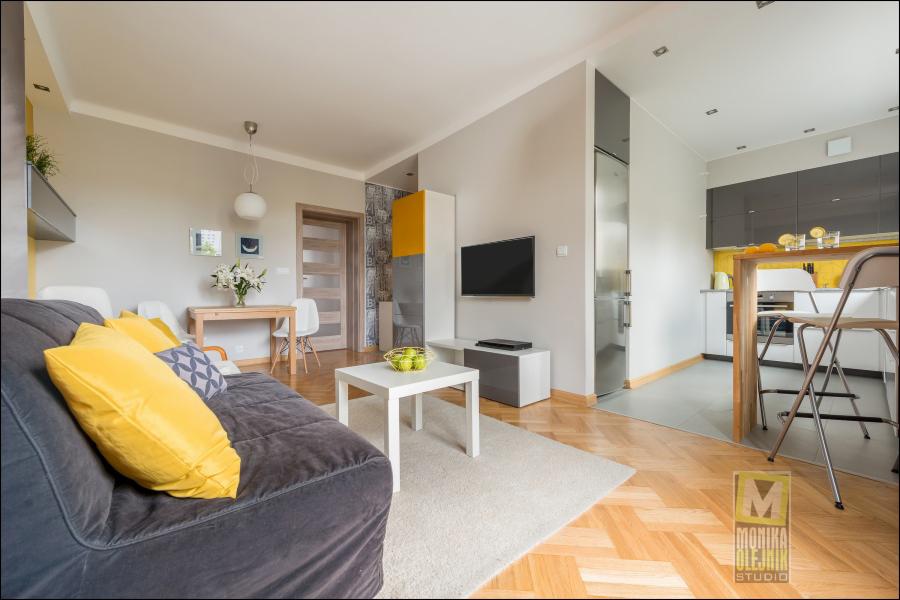 Jasne wnętrze mieszkania z szarą sofą z żółtymi poduszkami, białym stolikiem kawowym i widokiem na kuchnię z żółtymi akcentami, parkiet w jodełkę.