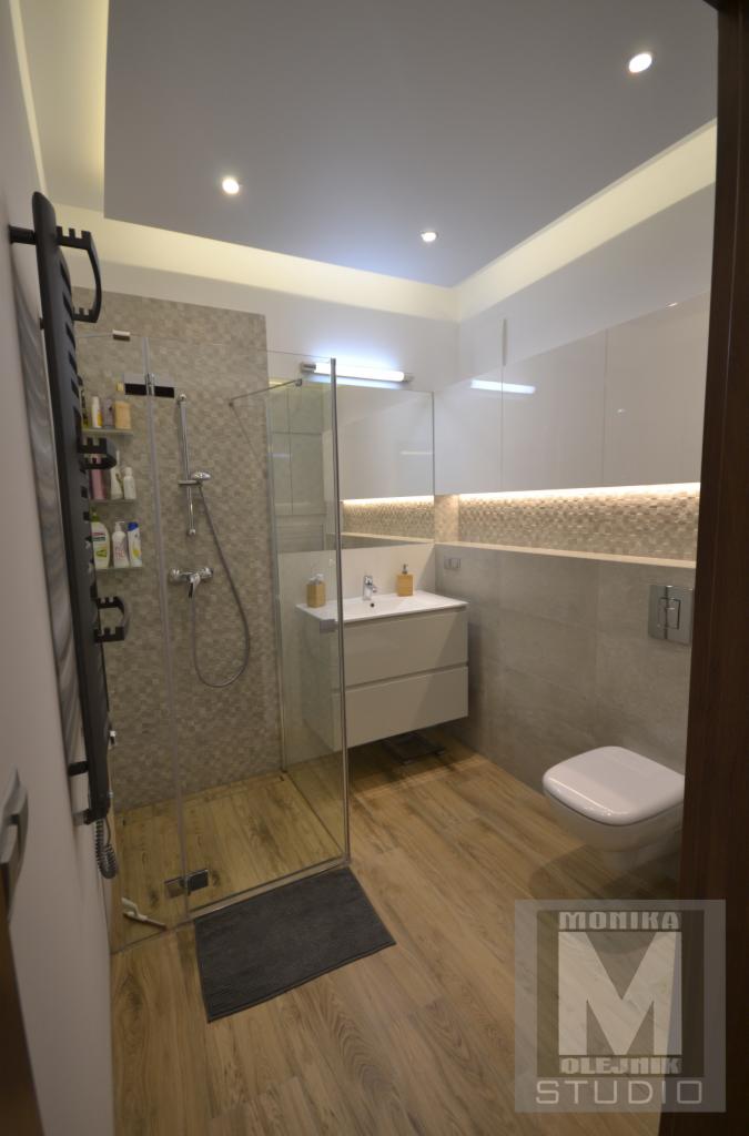 Nowoczesna łazienka z kabiną prysznicową typu walk-in, umywalką z szafką, podświetlanym lustrem i drewnopodobną podłogą, akcentowana szarym dywanikiem przy wejściu do kabiny.