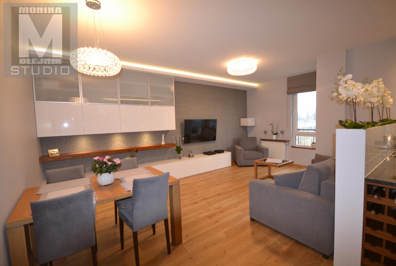 Nowoczesny salon z drewnianą podłogą, szarymi meblami, oświetleniem LED, stołem jadalnym z bukietem kwiatów i zabudową meblową z przeszklonymi szafkami.