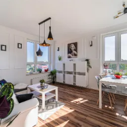 Jasne, przestronne wnętrze salonu z drewnianą podłogą, szarą sofą z kolorowymi poduszkami, białym stolikiem kawowym, białym regałem modułowym, stołem z czterema krzesłami i dużymi oknami z widokiem...