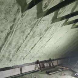 Poddasze z widoczną, nałożoną pianką izolacyjną, drewnianymi belkami stropowymi oraz rozłożoną drabiną i przewodami na podłodze.