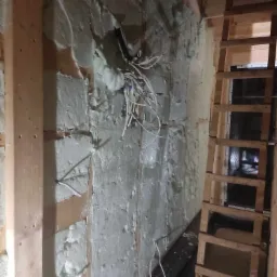 Ściana z drewnianą konstrukcją wypełniona białą pianką izolacyjną, widoczne przewody elektryczne i drewniana drabina w tle, oświetlenie robocze.