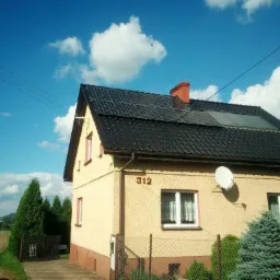 Dom jednorodzinny z ciemną dachówką, na której zamontowane są panele słoneczne i kolektory słoneczne, widoczny z poziomu gruntu w słoneczny dzień.