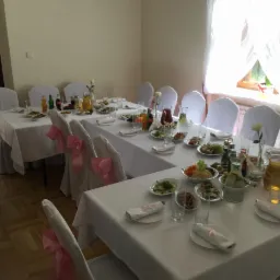 Uroczysta sala z długimi stołami nakrytymi białymi obrusami, zastawionymi talerzami z przystawkami i napojami, krzesła z białymi pokrowcami ozdobione różowymi kokardami, przygotowana na przyjęcie.