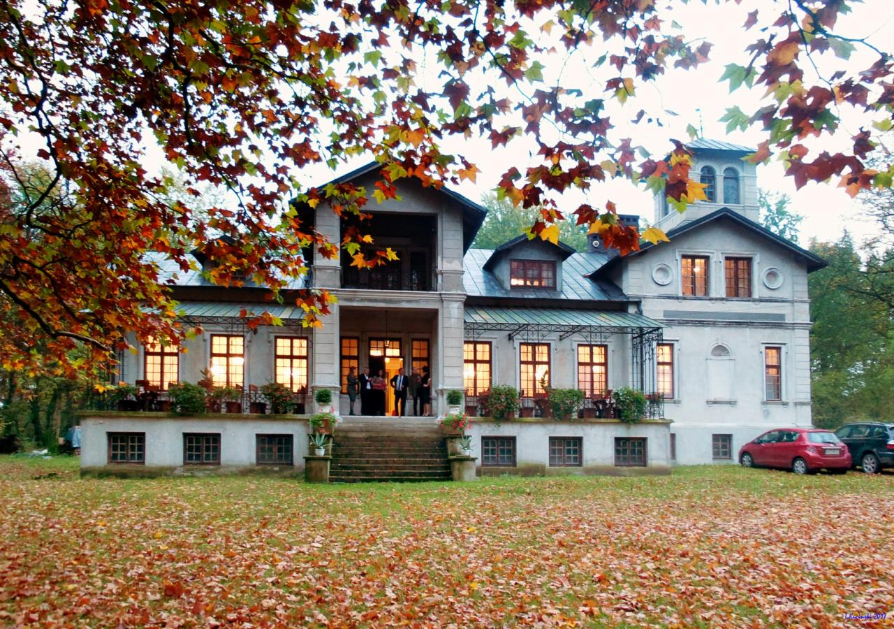 Dwupiętrowy budynek w stylu dworkowym z podświetlonymi oknami, przed budynkiem trawnik pokryty opadłymi liśćmi, w tle drzewa z jesiennymi liśćmi, na schodach do wejścia stoją ludzie.