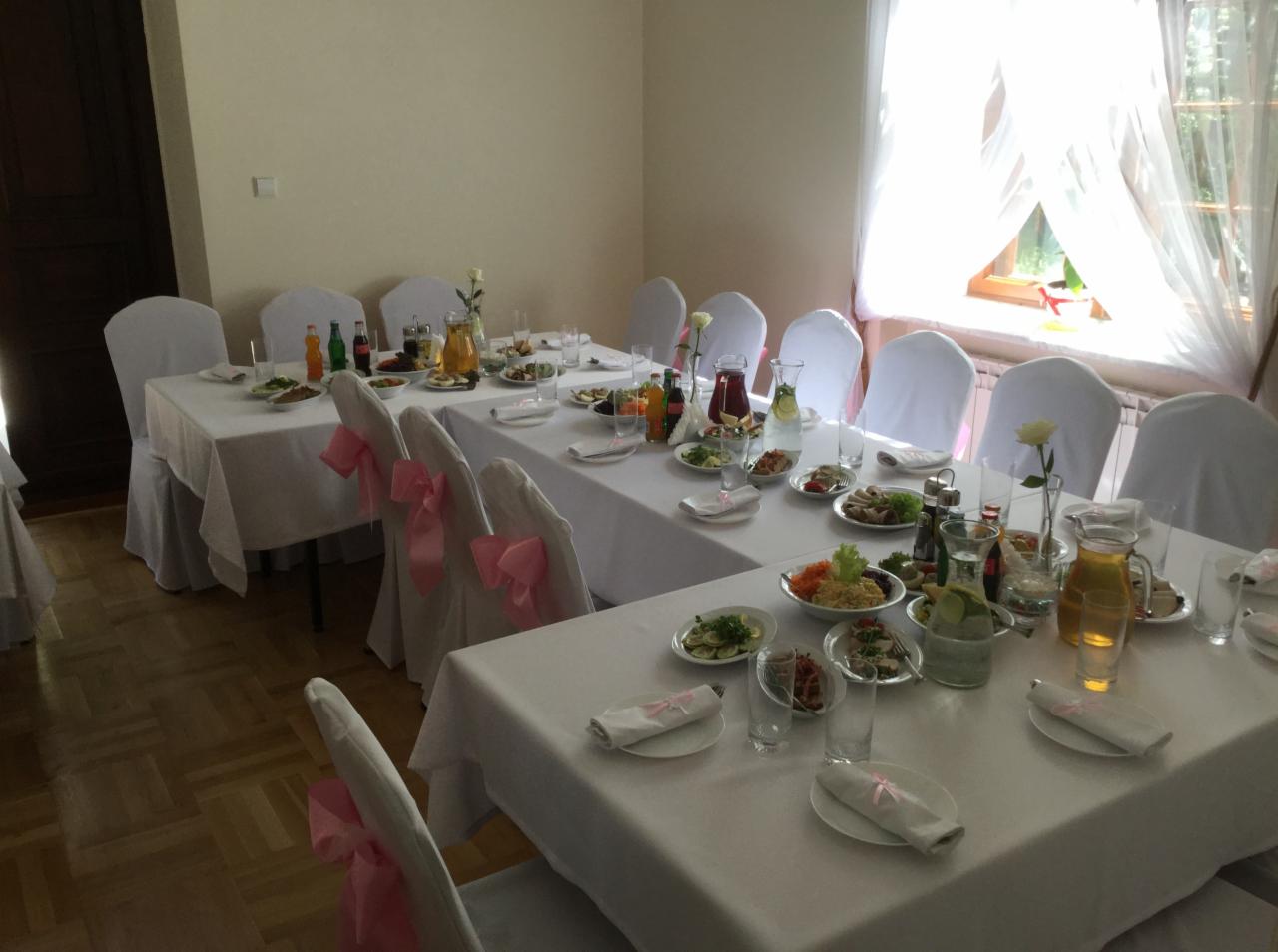 Uroczysta sala z długimi stołami nakrytymi białymi obrusami, zastawionymi talerzami z przystawkami i napojami, krzesła z białymi pokrowcami ozdobione różowymi kokardami, przygotowana na przyjęcie.