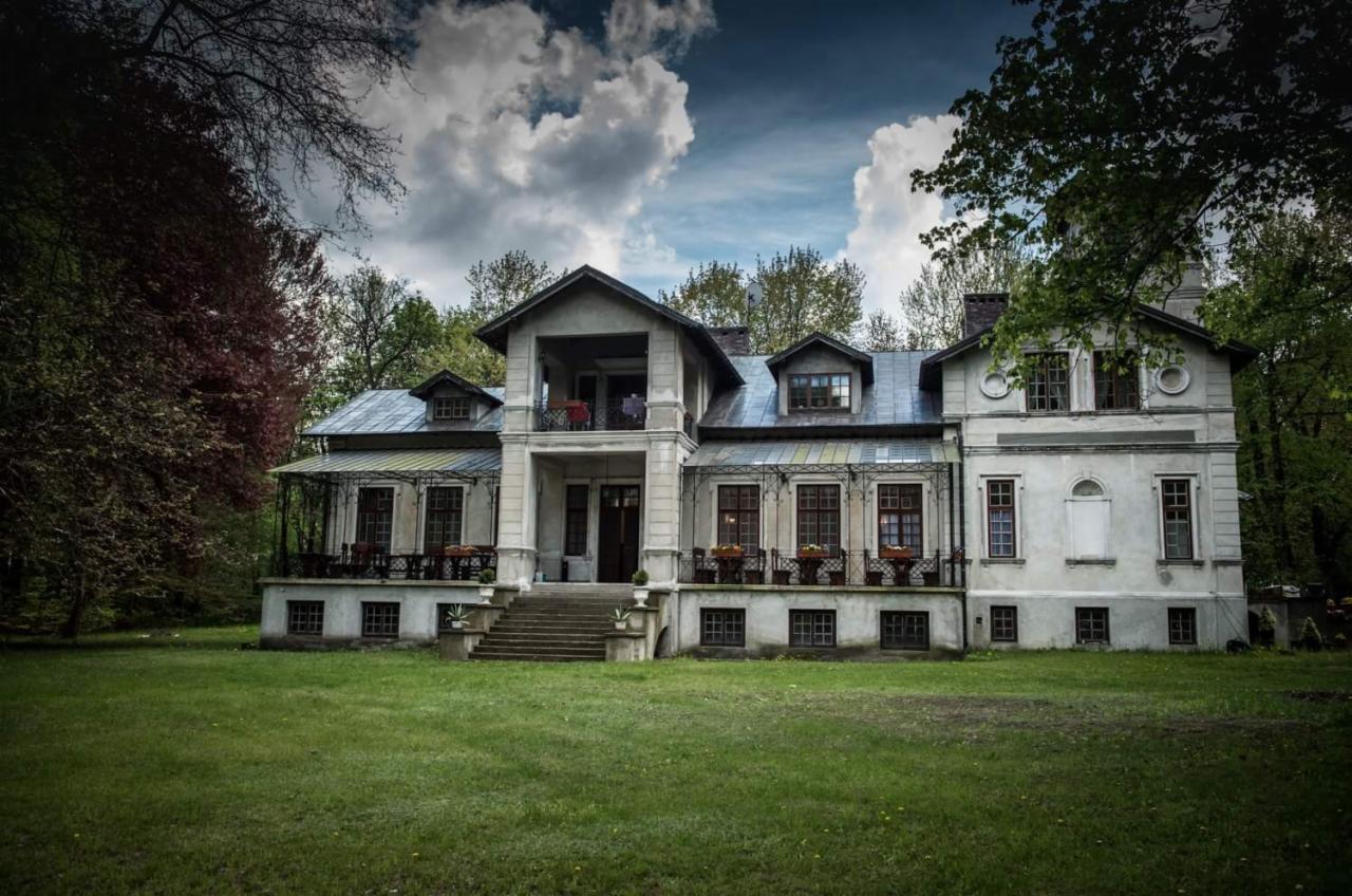 Dwupiętrowy, jasny dwór z kolumnami i balkonem, otoczony zielonym trawnikiem i drzewami w pochmurny dzień.